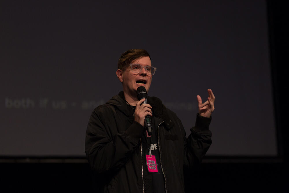 A person with short hair and glasses speaking into a microphone. Their left hand is holding the microphone and their right hand is up in the air gesturing. 