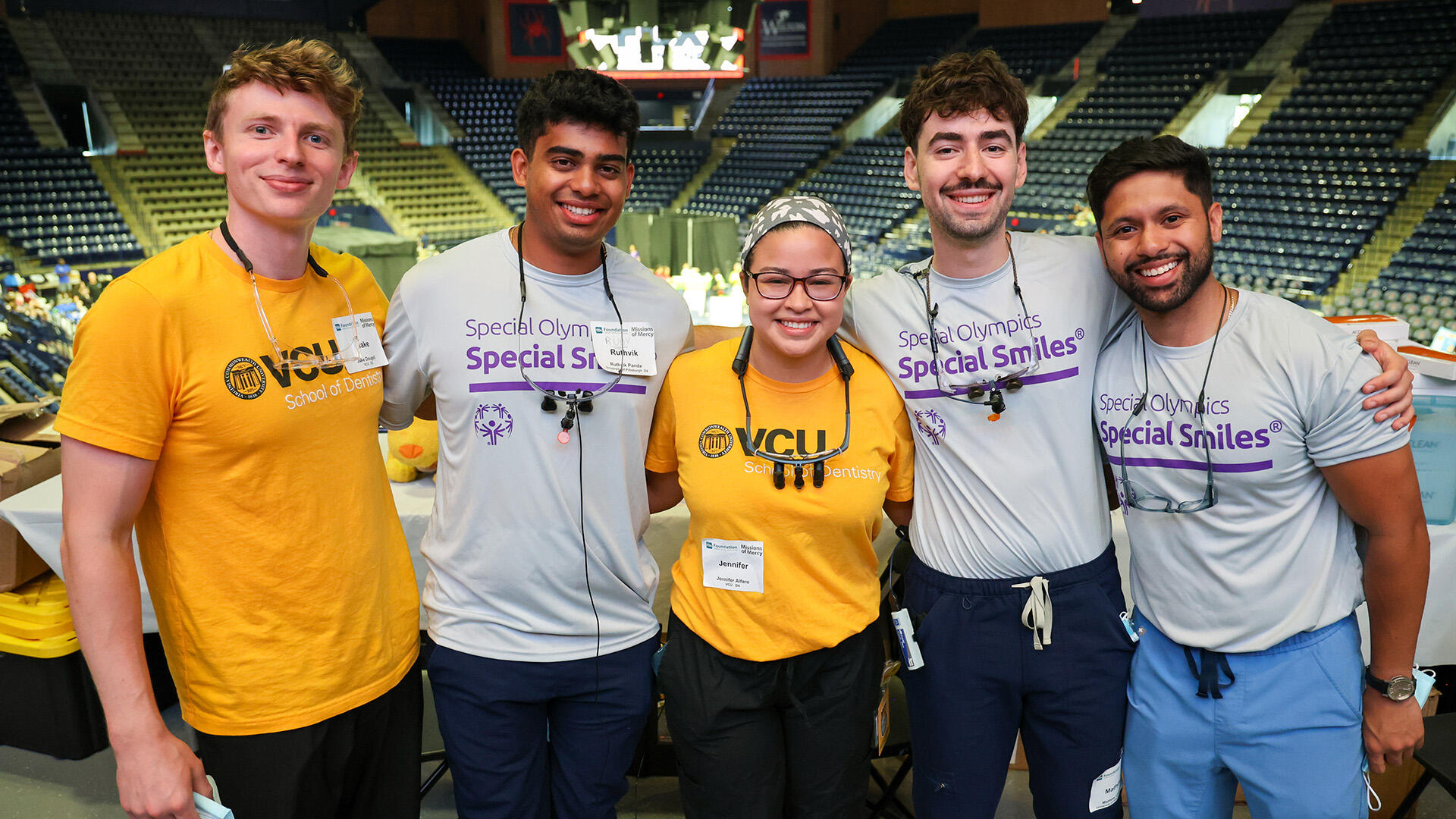 A group photo of five people standing in a row with their arms behind each other's backs. The person on the far left and the person in the middle are wearing yellow t-shirts that say \"VCU School of Dentistry\" in black and white text. The other three people are wearing gray sweatshirts that say \"Special Olympics Special Smiles\" in purple text. 