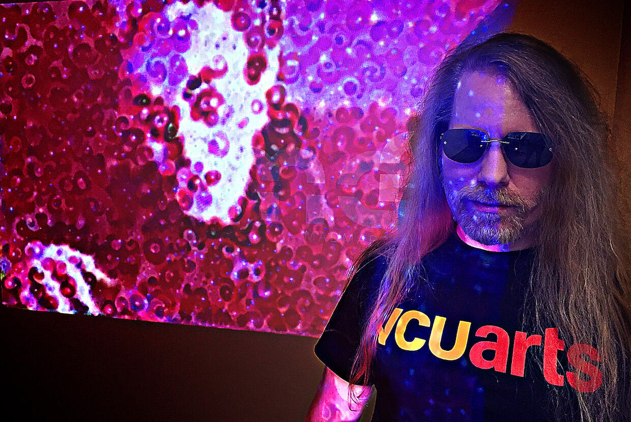 Eric Millikin, wearing a vcu arts t-shirt and sunglasses, stands in front of a video artistic projection of the character Nosferatu.