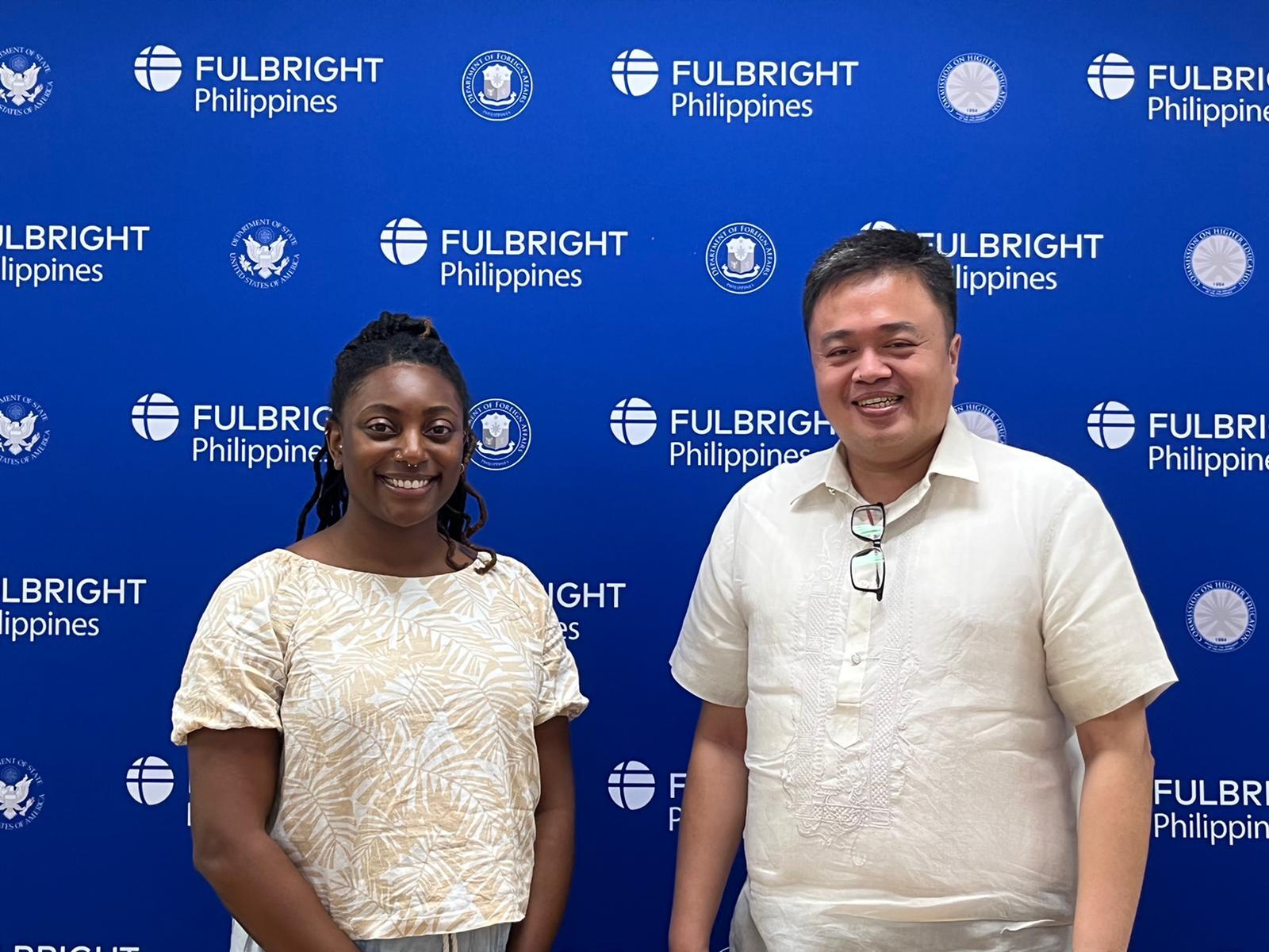 A woman standing to the right of a man in front of a blue wall with the phrase \"Fulbright Philippines\" written all over it. 