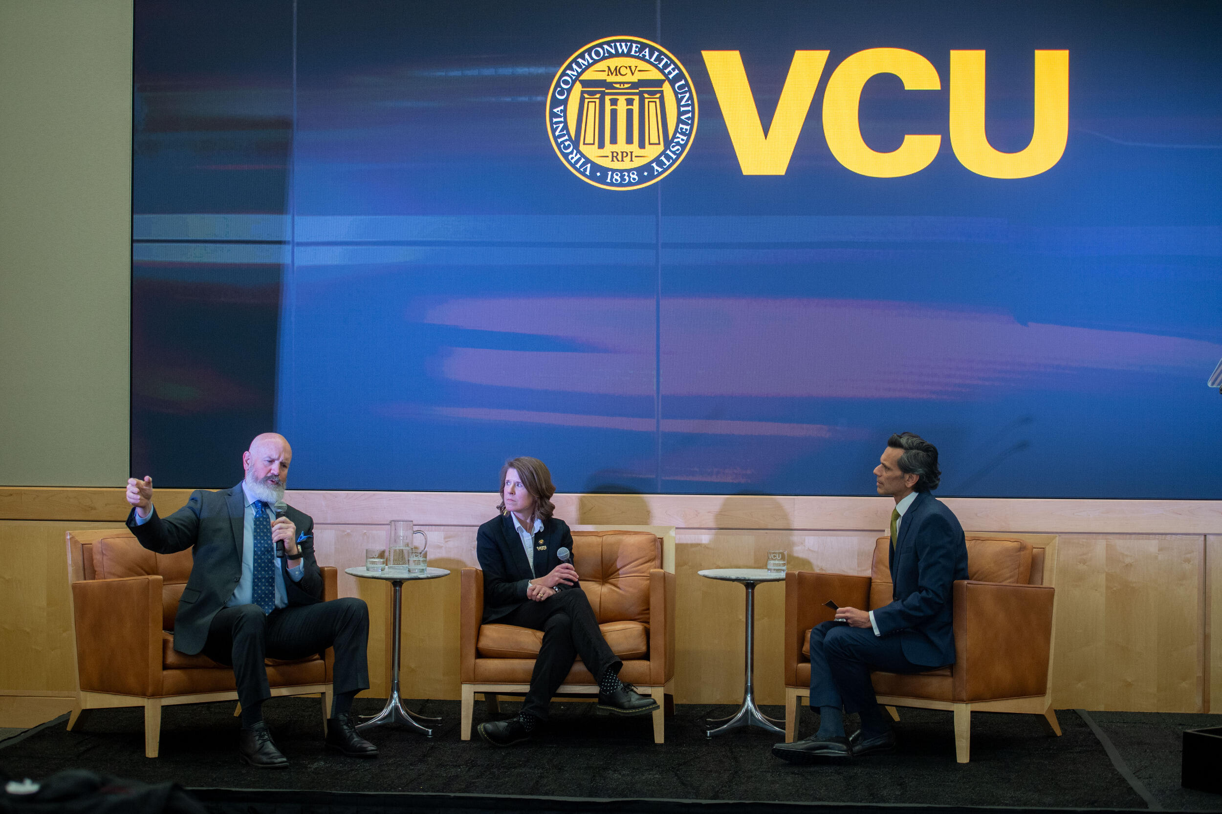 Three people sit in chairs on a stage. The man sitting on the left is gesturing and speaking.