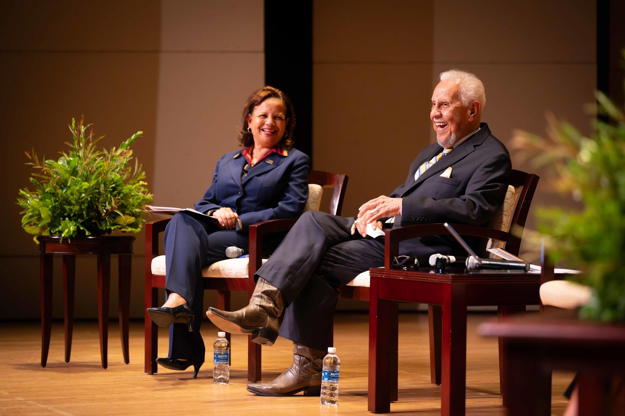 A photo of a man and a woman sitting in chairs on a stage. 
