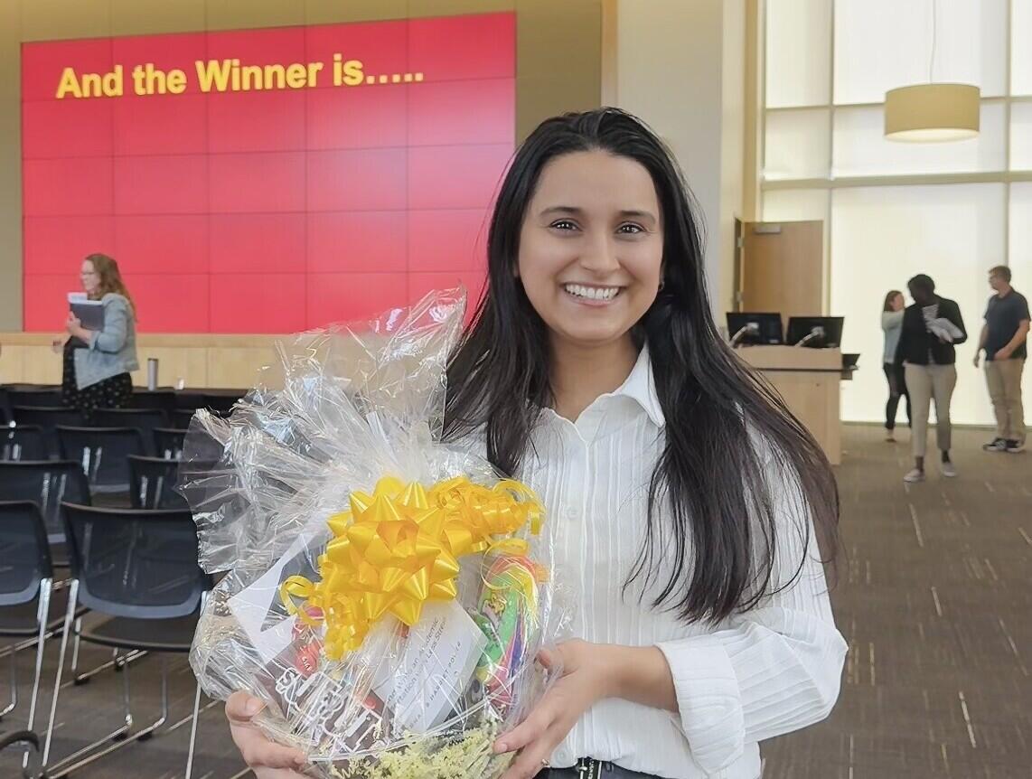 A photo of a woman from the knees up holding a gift basket with a yellow ribbon. Behind her is a red screen with yellow letters that says \"And the Winner is...\"