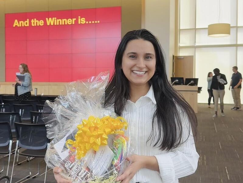 A photo of a woman from the knees up holding a gift basket with a yellow ribbon. Behind her is a red screen with yellow letters that says \"And the Winner is...\"