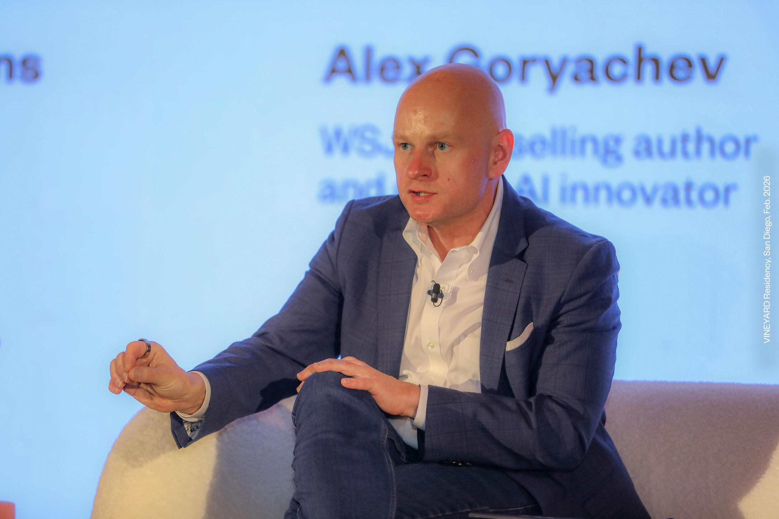 A man in a suit sits in a chair and gestures while he speaks.