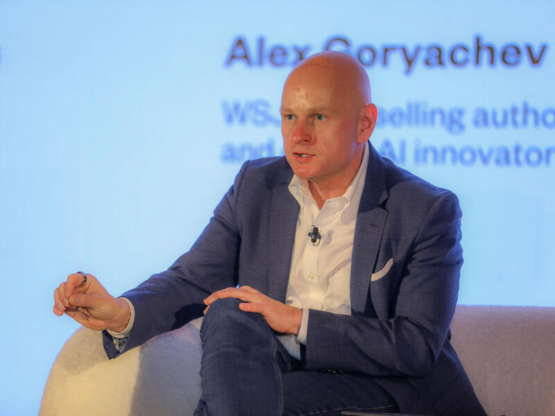 A man in a suit sits in a chair and gestures while he speaks.
