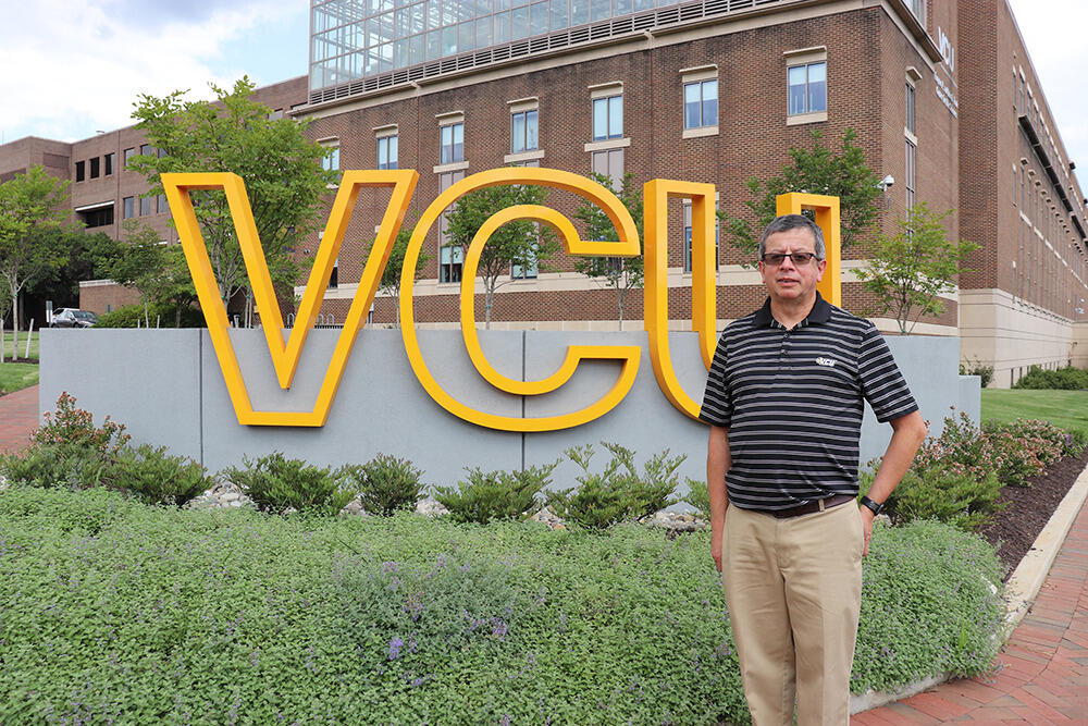 A man wearing a black collared shirt standing in front of a sign that says \"V C U\" 