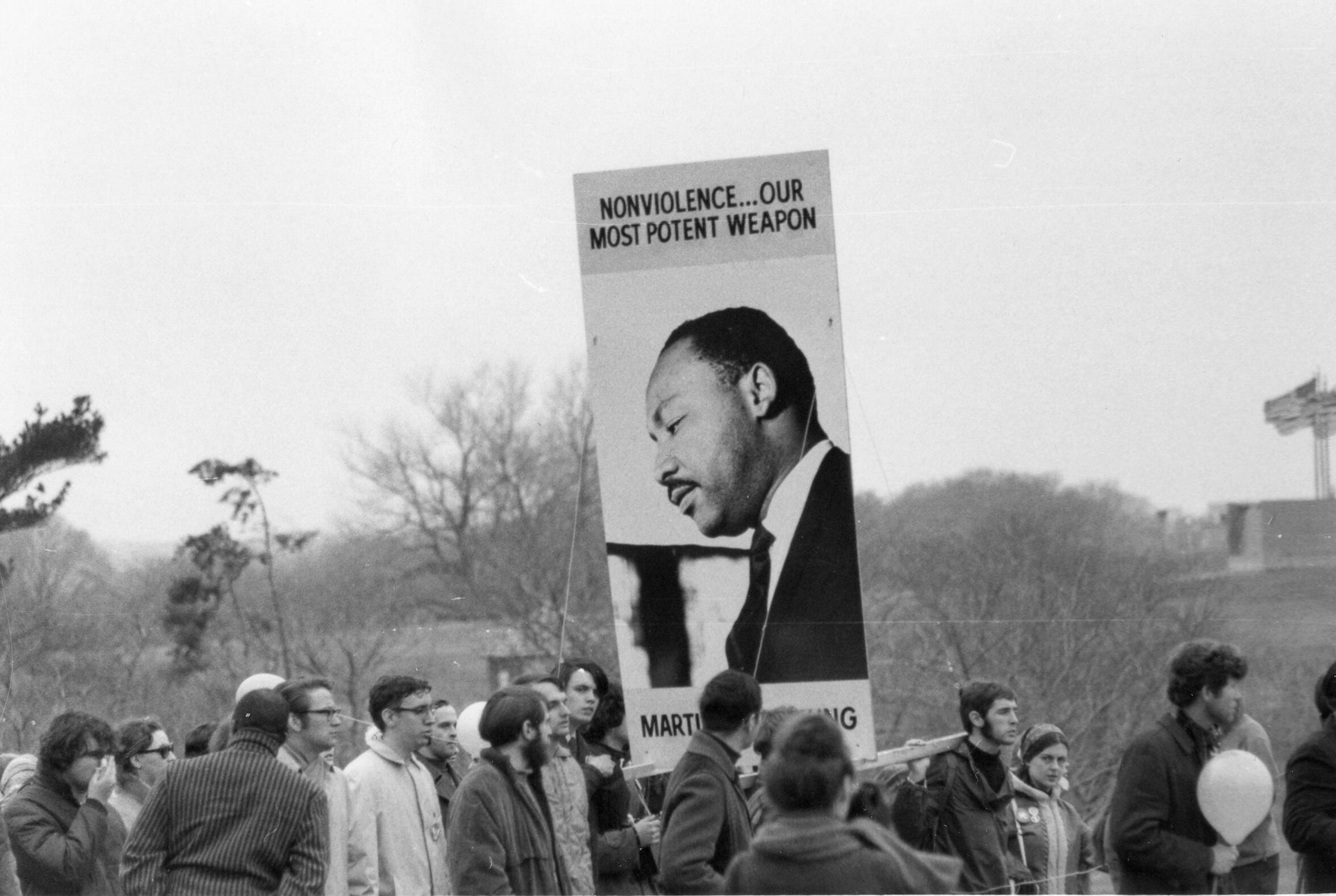 A black and whie photo of a crowd of people. In the middle of the crowd is a sign with a photo of Martin Luther King Jr. that says \"NONVIOLENCE.... OUR MOSTPOTENT WEAPON\" 