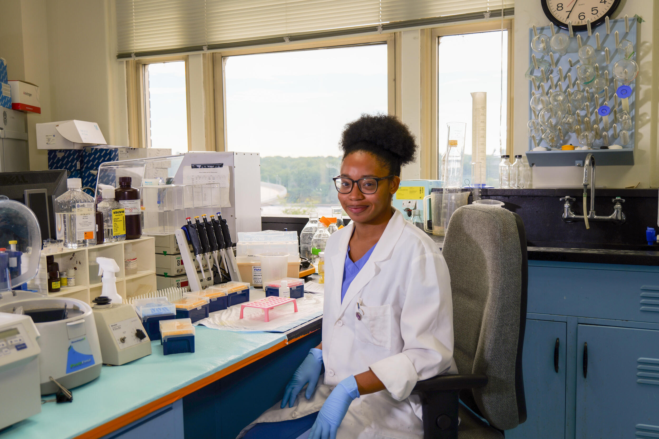 A photo of a woman wearing a white medical coat, blue scrubs, blue latex globes, and safety glasses sitting in a medical lab.