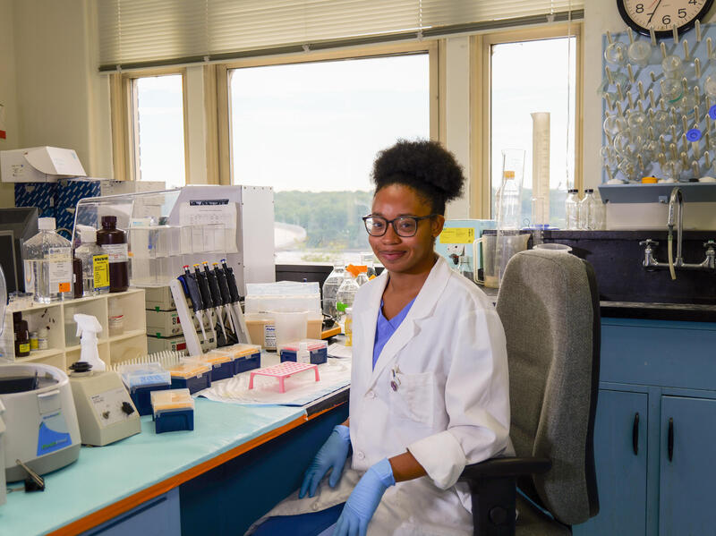 A photo of a woman wearing a white medical coat, blue scrubs, blue latex globes, and safety glasses sitting in a medical lab. 