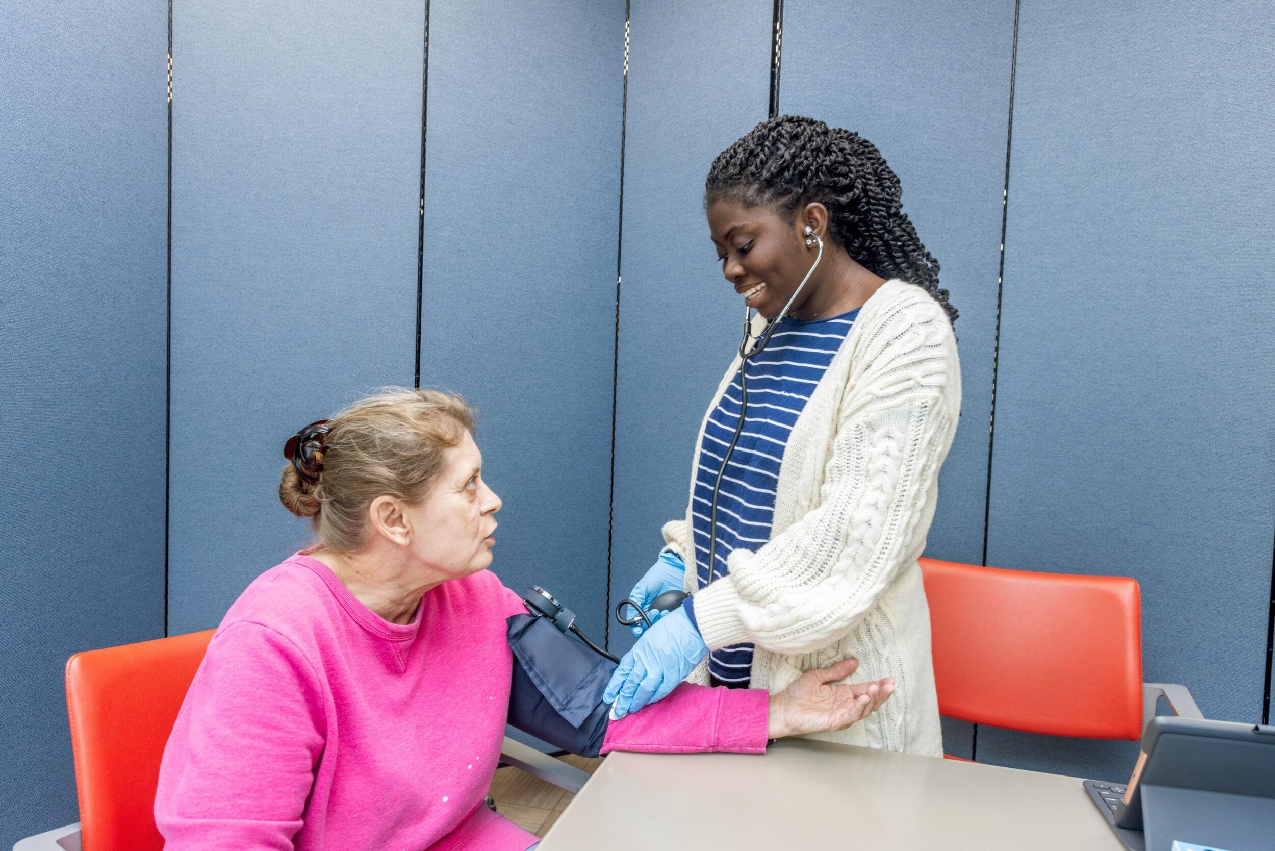 A photo of a woman wearing a white lap coat listening to the heartbeat of a woman sitting down. 