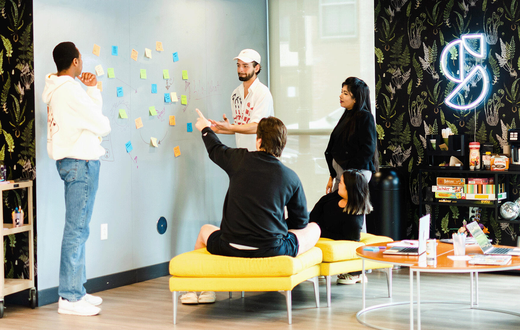 A photo of three people standing and one person sitting in front of a wall with sticky notes on it. 