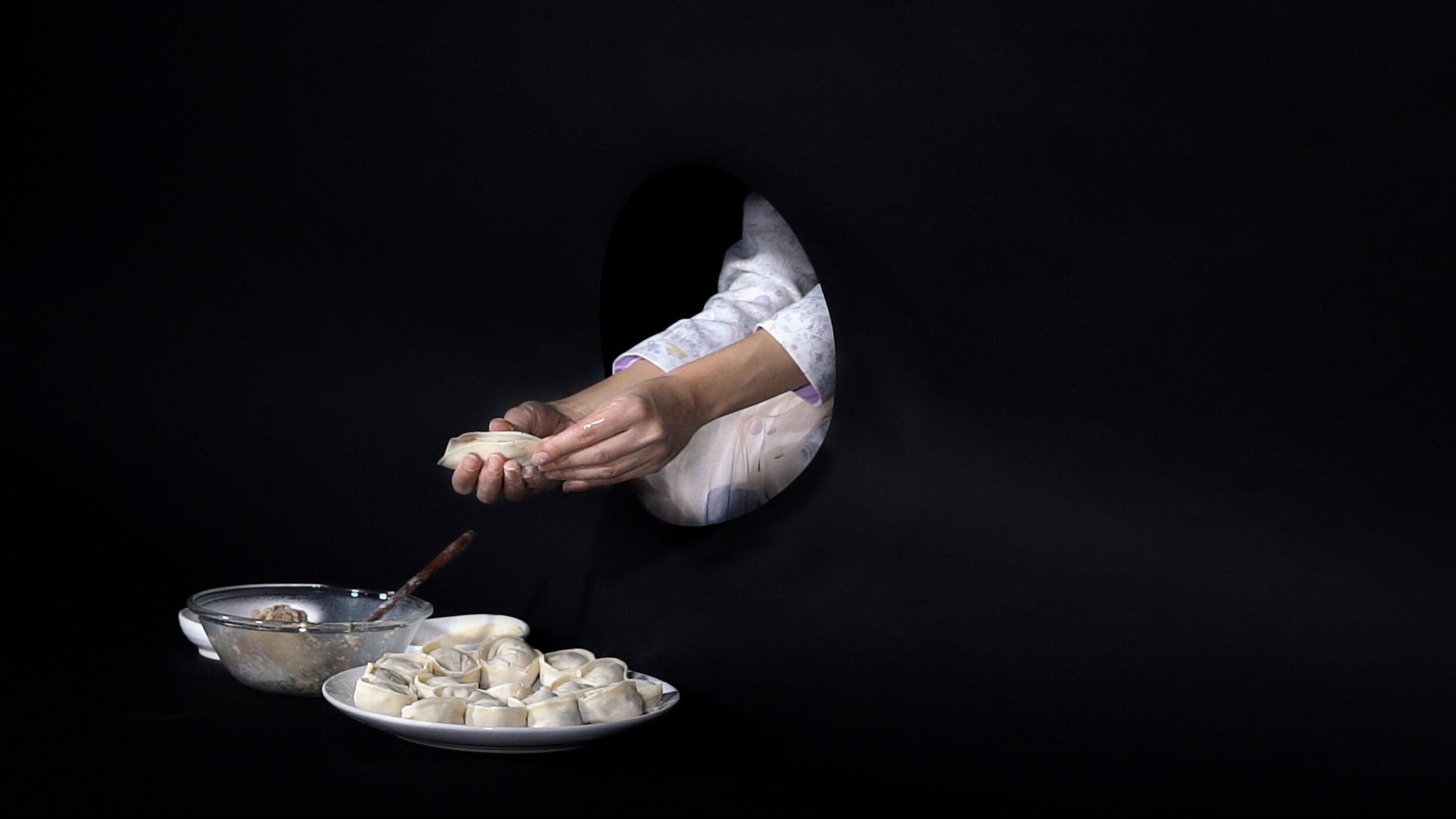 An image from Cecilia Hankyeol Kim's \"Performed Labor.\" Two hands filling a dumpling in front of a bowl of filling which is next to a plate of prepared dumplings. 