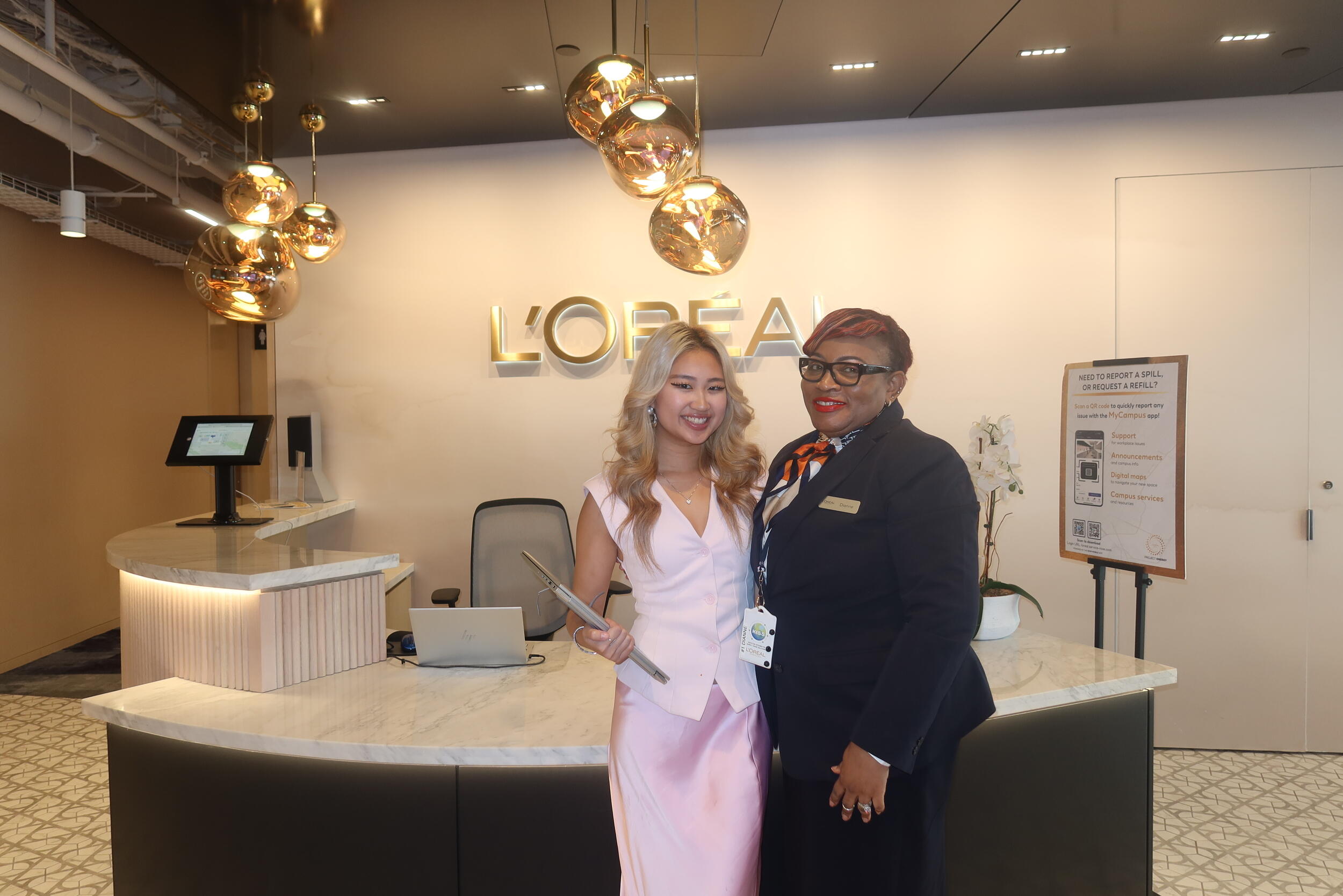 A photo of two women standing in front of a white secretary desk. Behind the desk is a gold sign that says \"L'OREAL\"