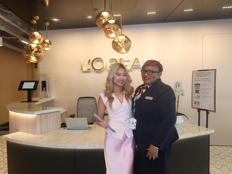 A photo of two women standing in front of a white secretary desk. Behind the desk is a gold sign that says \"L'OREAL\"