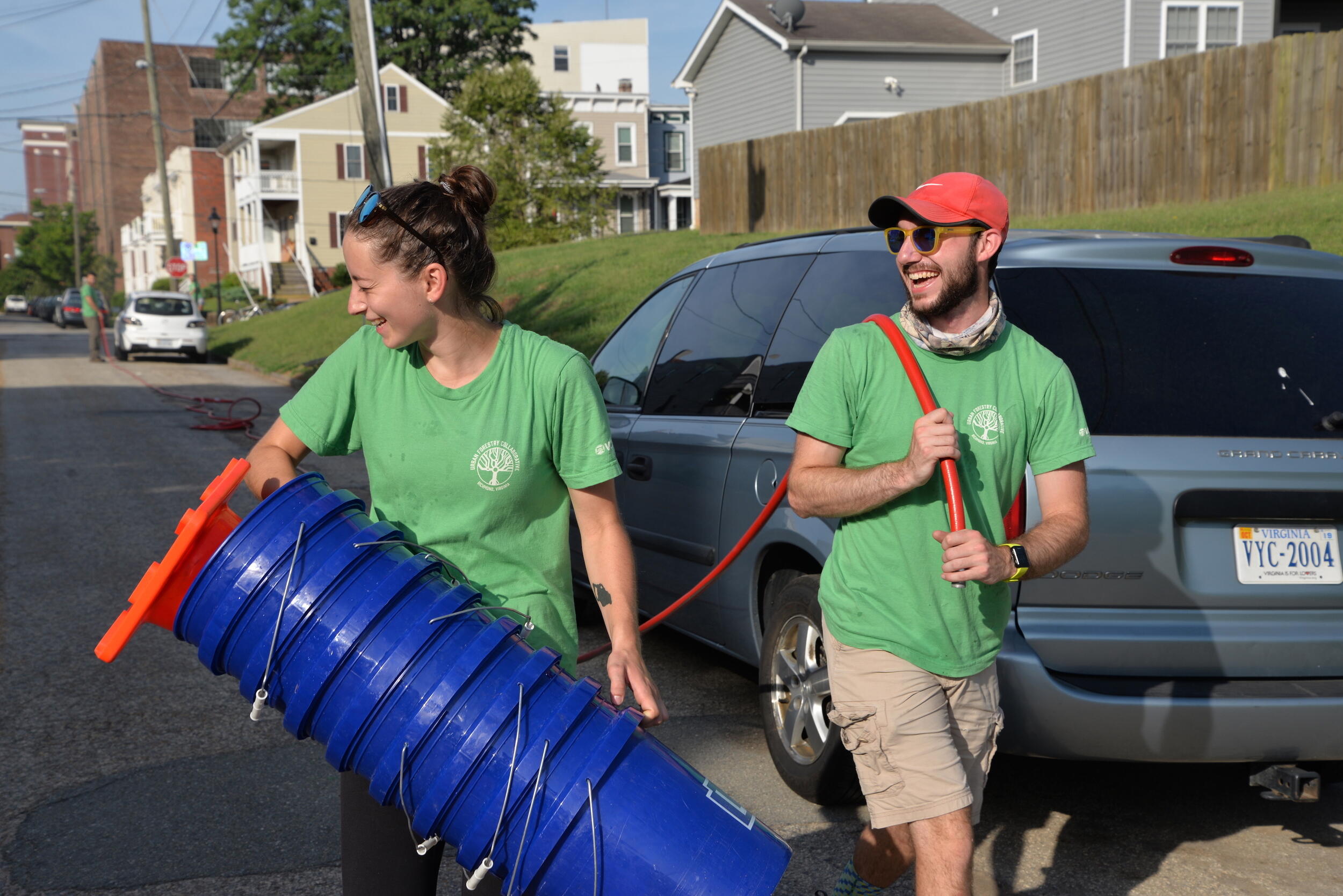 Two laughing students — one holding a stack of blue buckets, left, and another pulling a hose — are walking down a street.