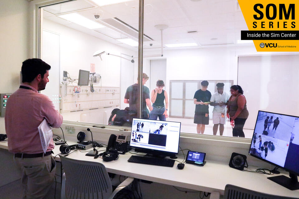 A photo of a man standing behind a desk looking through a glass window. He is watching five people interacting on the other side. 