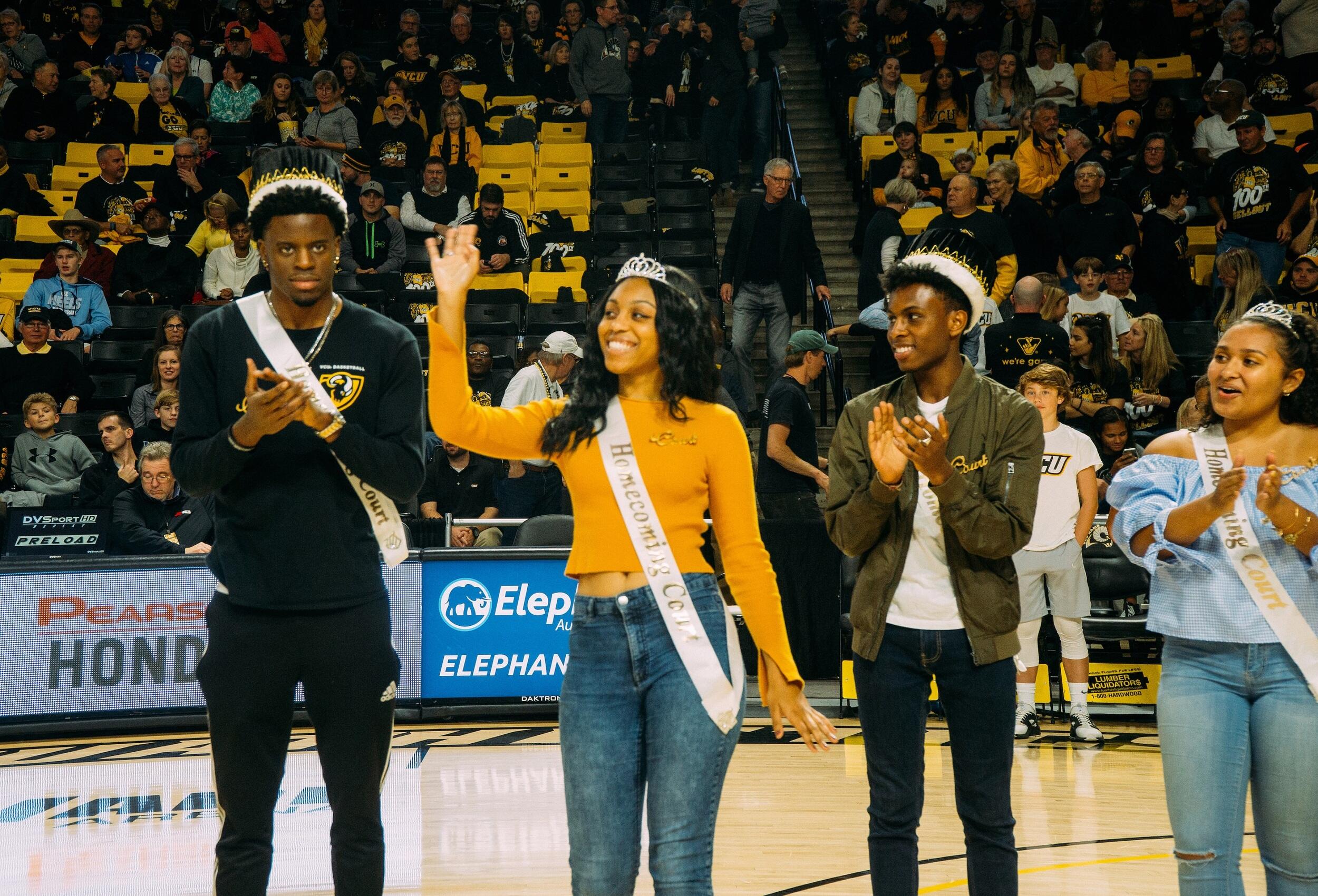 Two men and two women wearing crowns and sashes standing on a basketball court. 