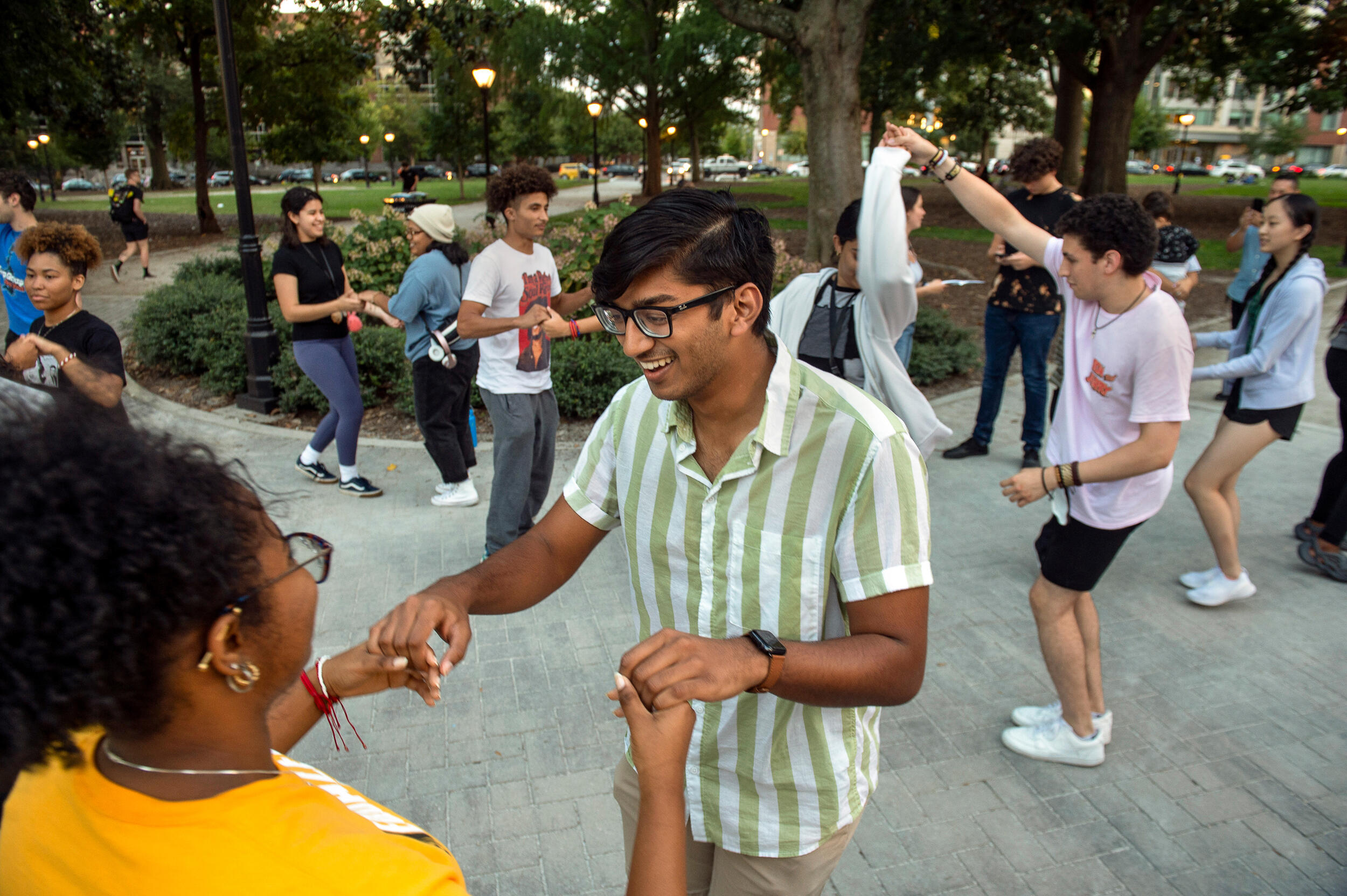 A student dances with another student while other students dance around a brick sidewalk next to a green space in Monroe Park.