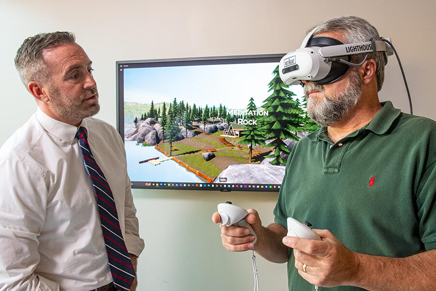 A photo of a man watching another man use a VR headset.