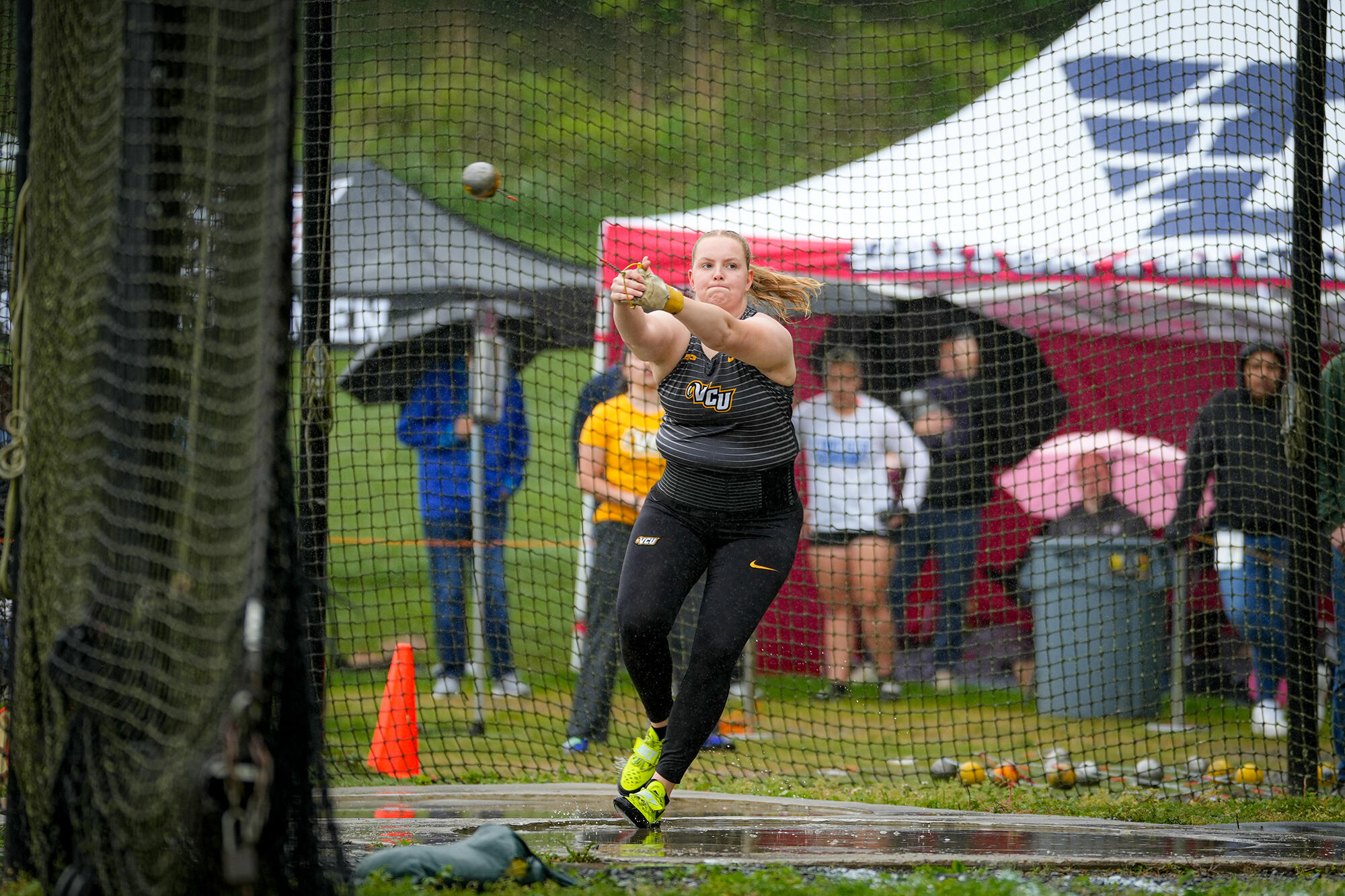 A photo of a woman jumping while holding her arms in front of herself. Behind the woman is a black netted fence with six people standing and sitting behind it. 