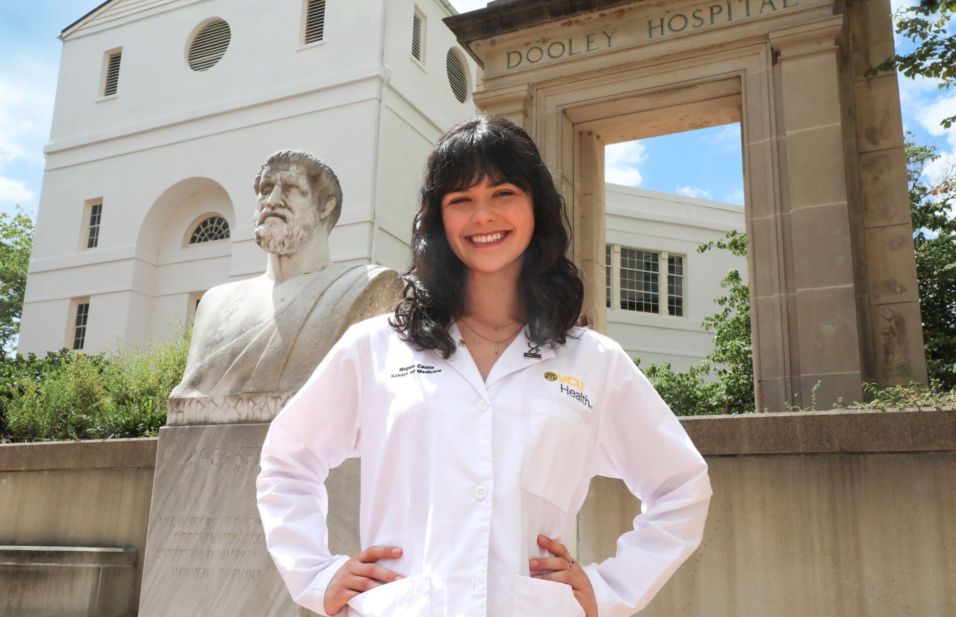 A photo of a woman wering a white medical coat from the waist up. She is standing with her hands on her hips and smiling. 