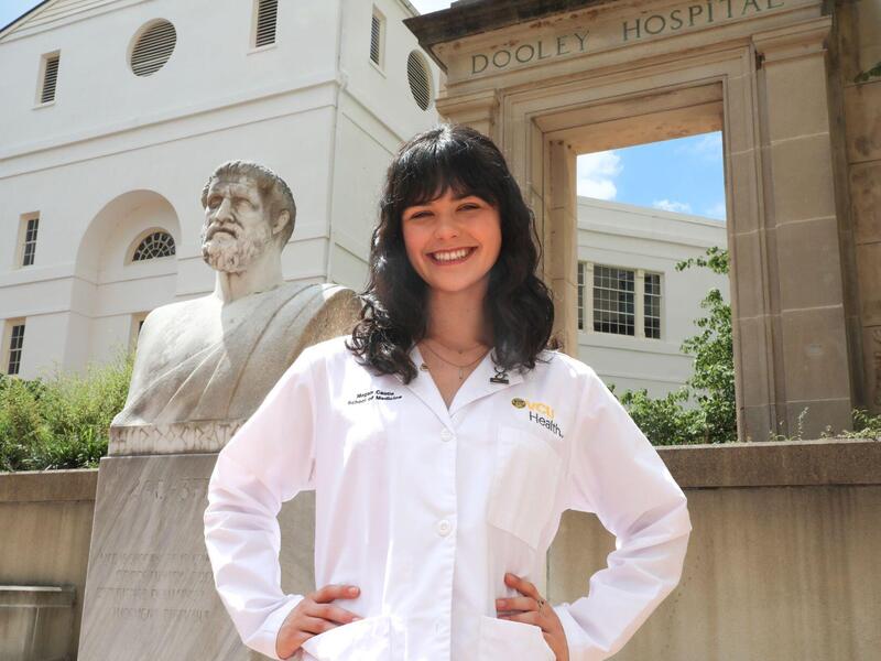 A photo of a woman wering a white medical coat from the waist up. She is standing with her hands on her hips and smiling. 