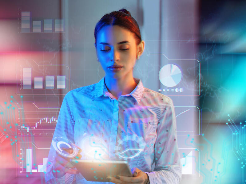 A photo of a woman using a tablet. Around the woman are digital illustrations of sets of data. 