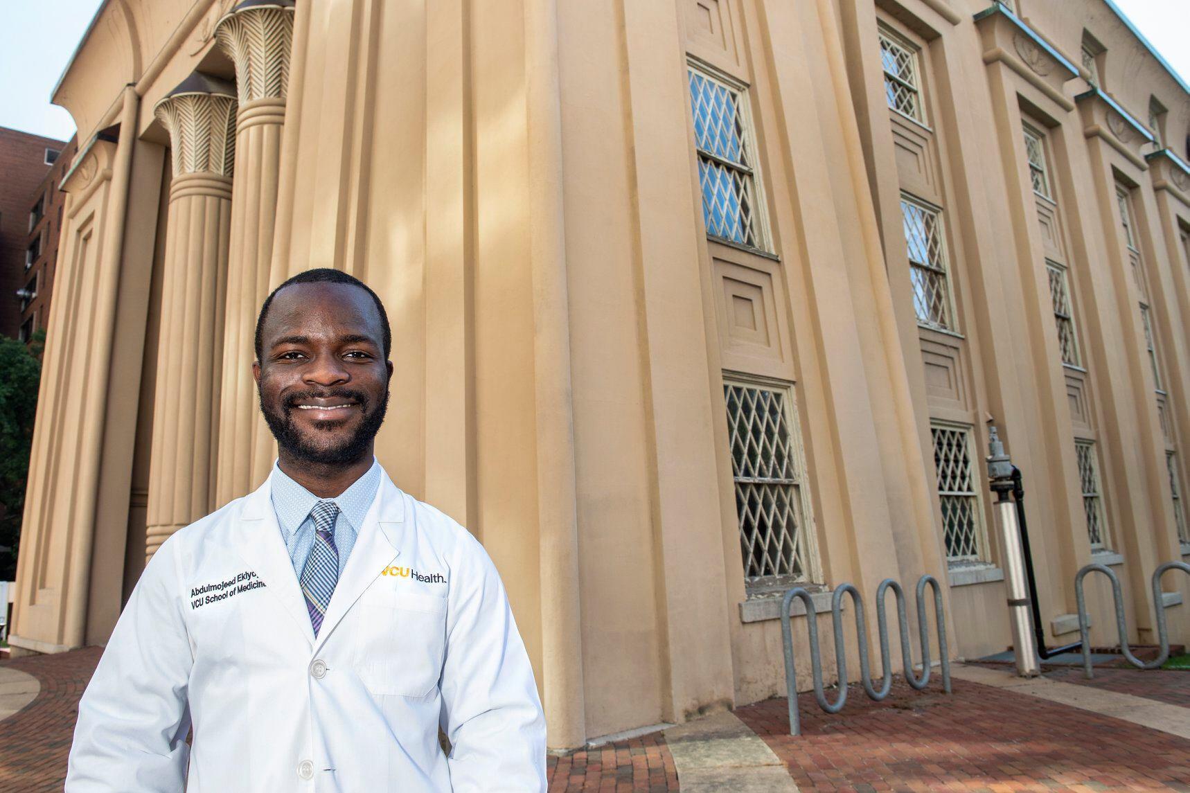 Abdulmojeed Ekiyoyo stands in front of the Egyptian Building on V C U's Medical College of Virginia campus. The right side of his jacket says Abdulmojeed Ekiyoyo, V C U School of Medicine. The left side of his jacket says V C U Health.