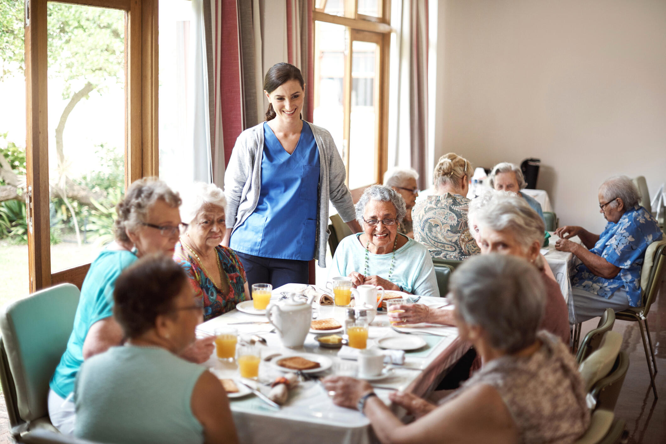 A dining room with two tables that have older people eating at them. There is a younger woman wearing a nurse's scrubs watching the people eat and smiling. 