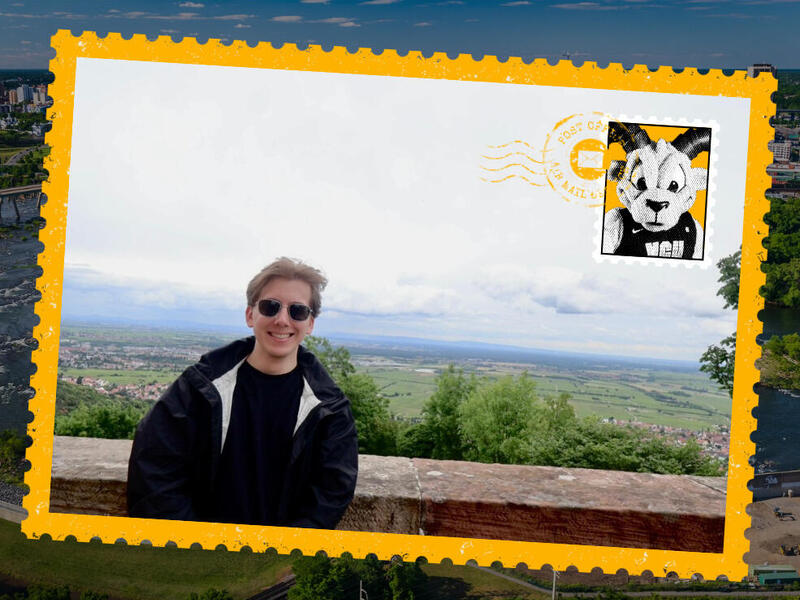 A photo of a postcard with a picture of a man standing in front of a short stone wall. Behind him are fields and a town. 