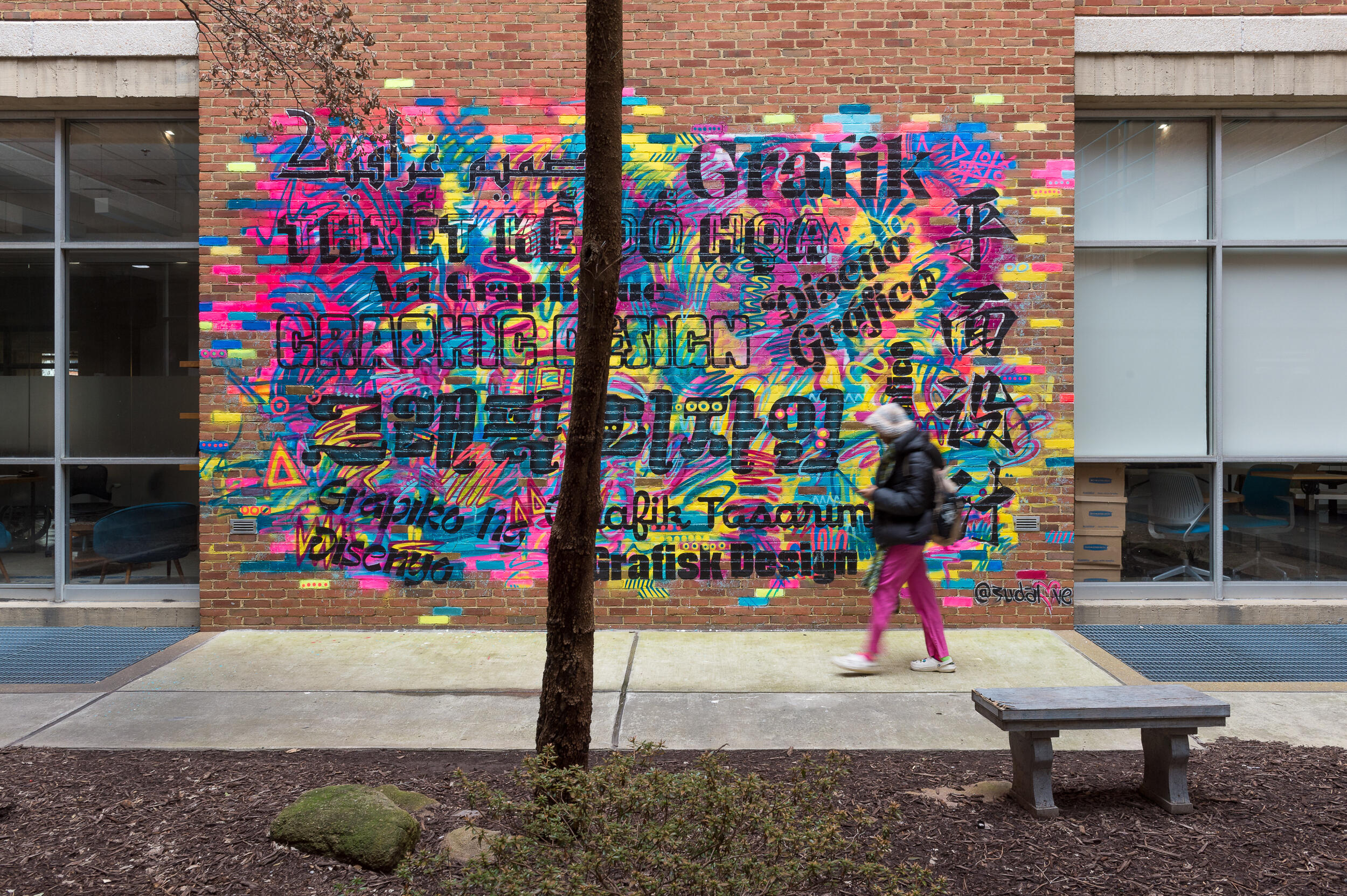 A student walks along a city sidewalk past the Pollak Building's brightly colored mural with the words \"graphic design\" translated into different languages.