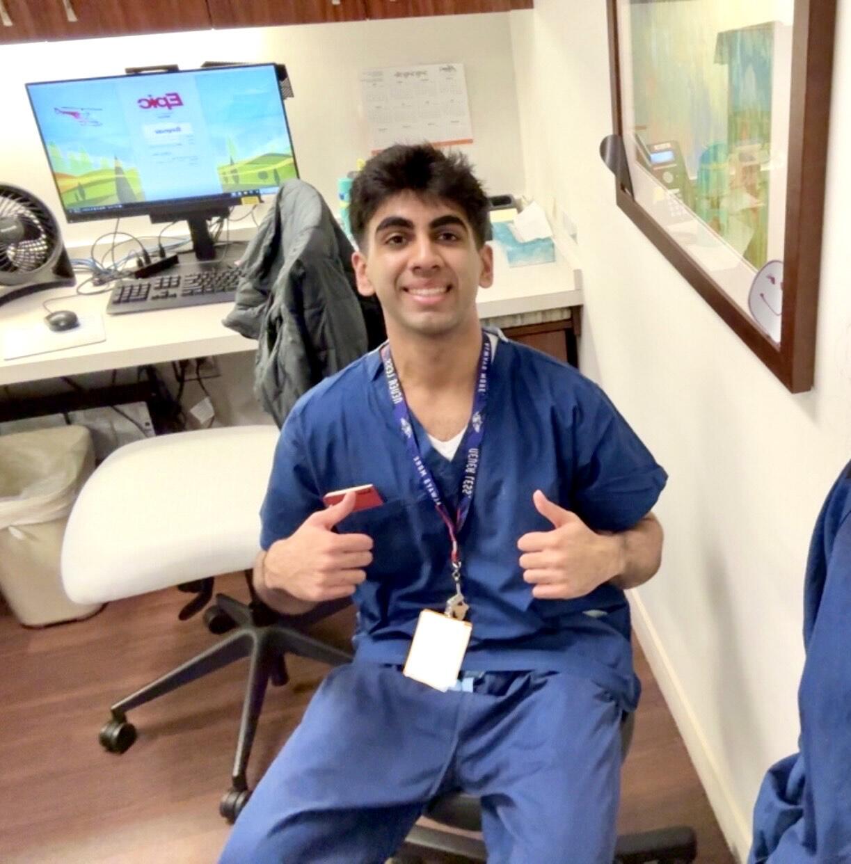 A man in blue hospital scrubs gives thumbs up, sitting in front of a computer screen.