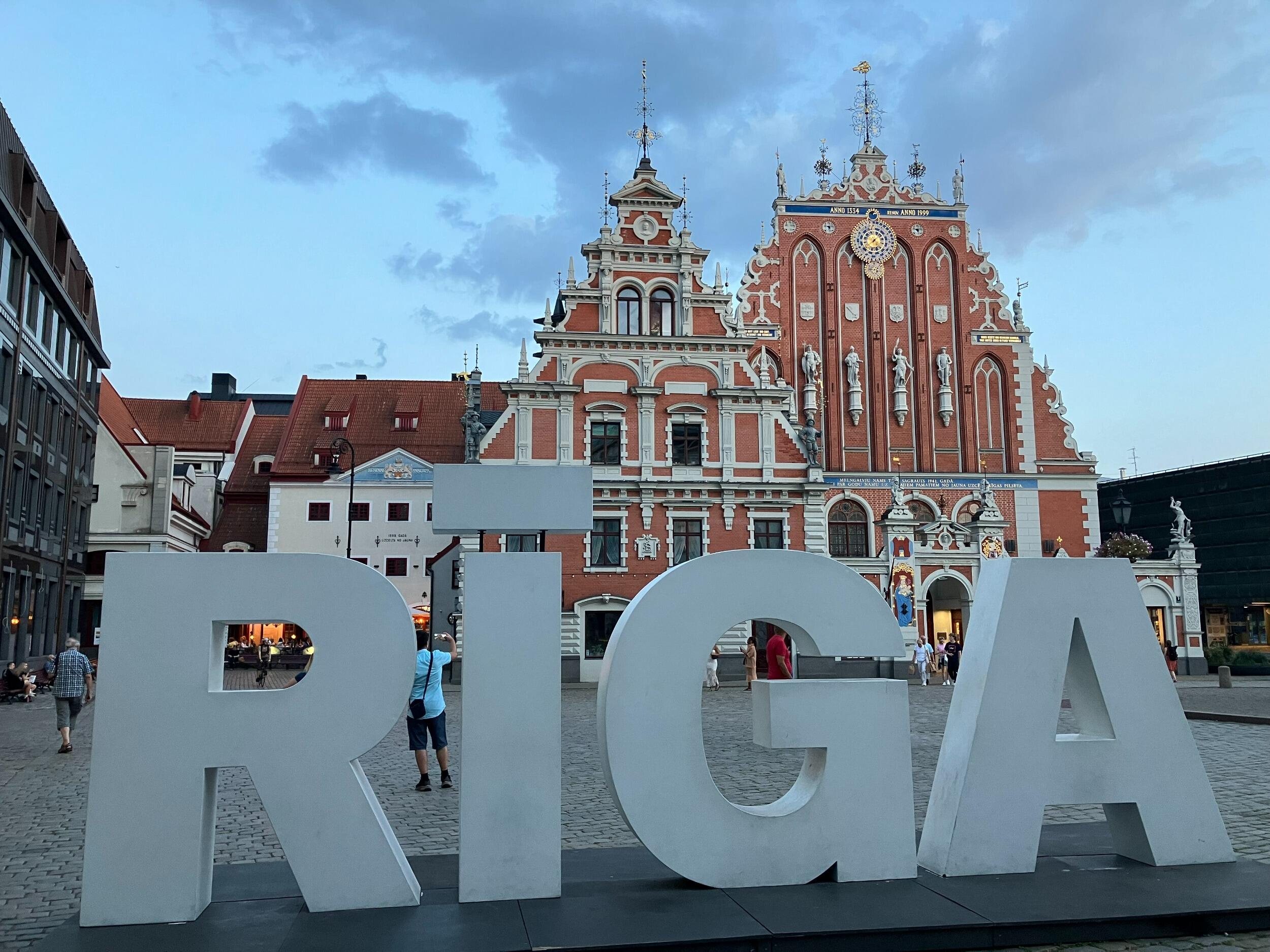 A photo of a sign that says \"RIGA\" in large white letters. The sign is in front of large old buildings in a town center. 