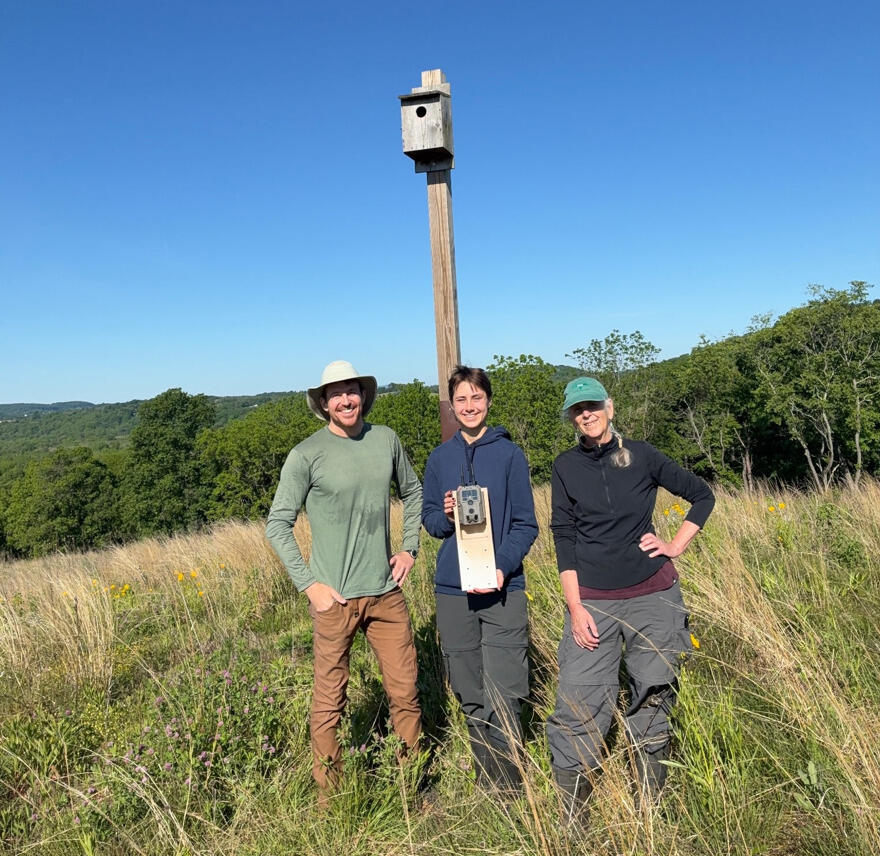 A photo of three people standing in front of a bird house set up in a field of tall grass. 