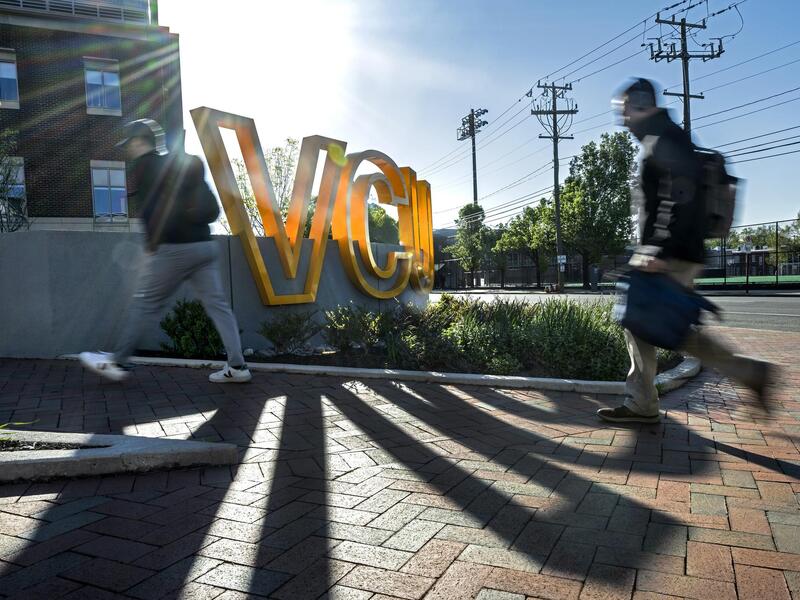 A photo of two people walking on a brick sidewalk. They are passing a sign that spells out \"VCU\" in giant yellow letters.