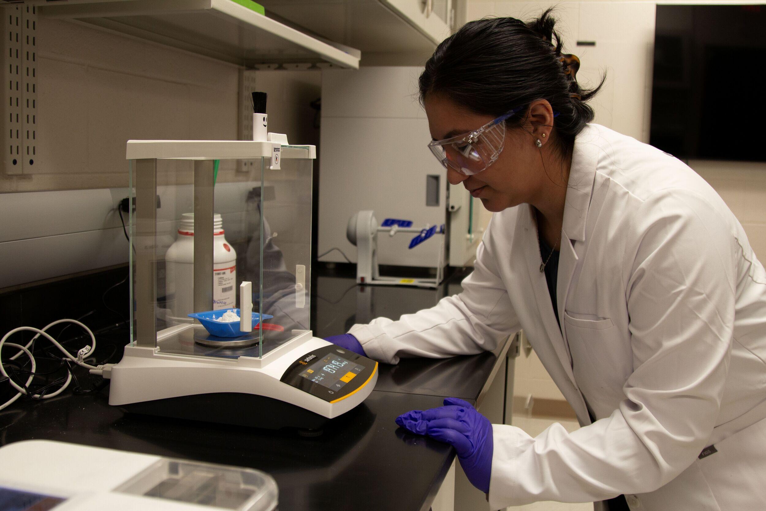 A woman in a lab coat and safety goggles leans over and looks closely at lab equipment.