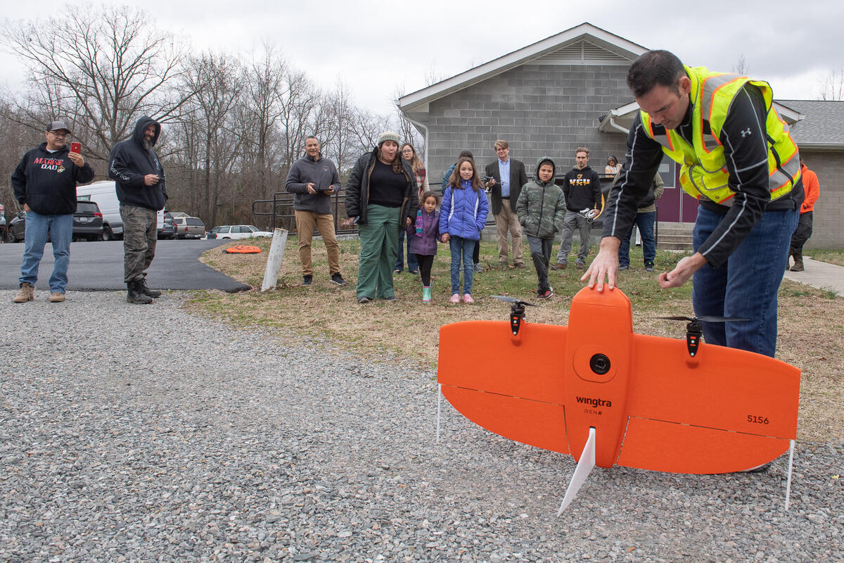 A photo of a man holding a large orange drone up as it balances on the ground. Behind him is a crowd of people watching him. 