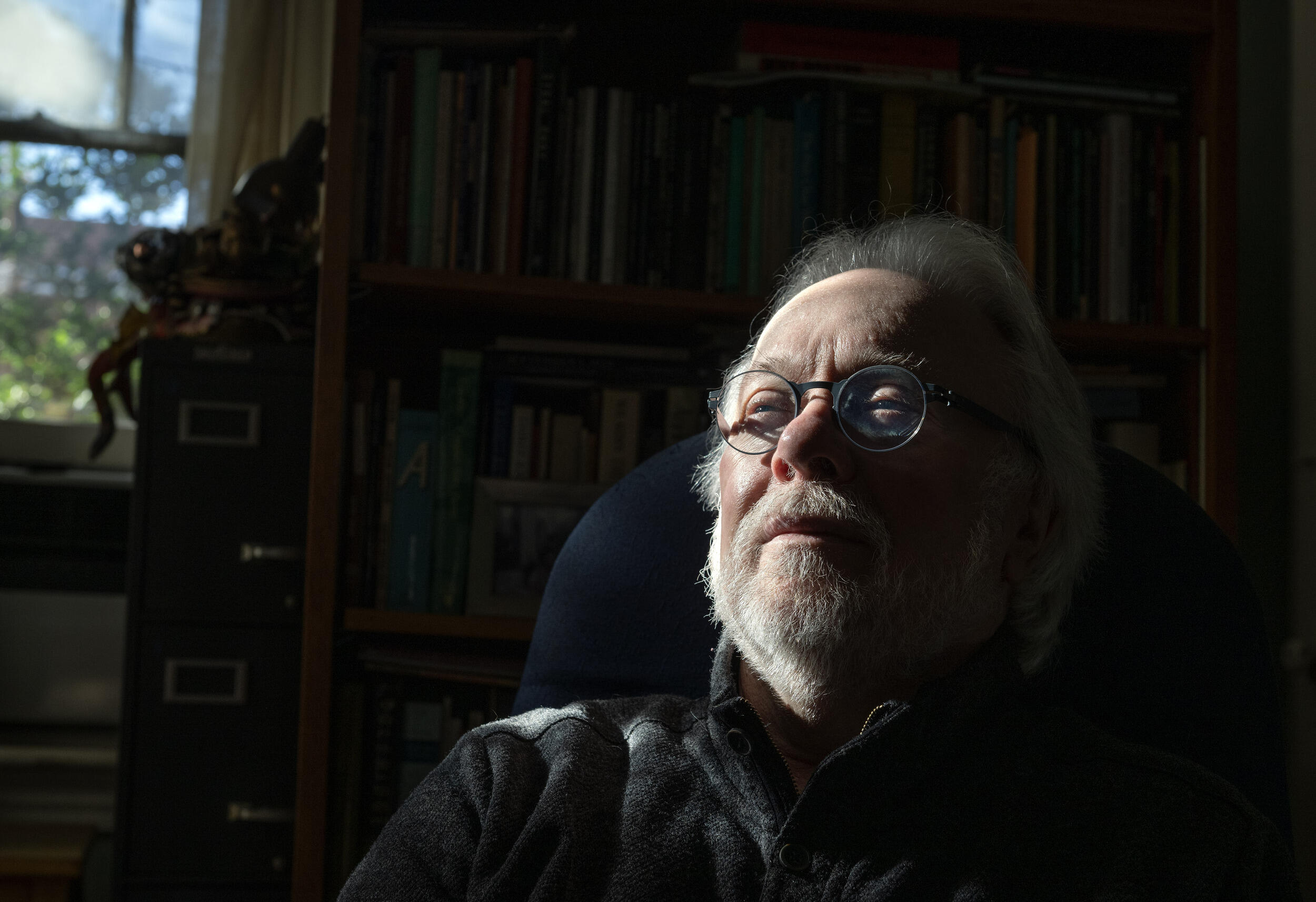 A photo of a man sitting in a chair from the shoulders up. Behind him is a book case and a window. 