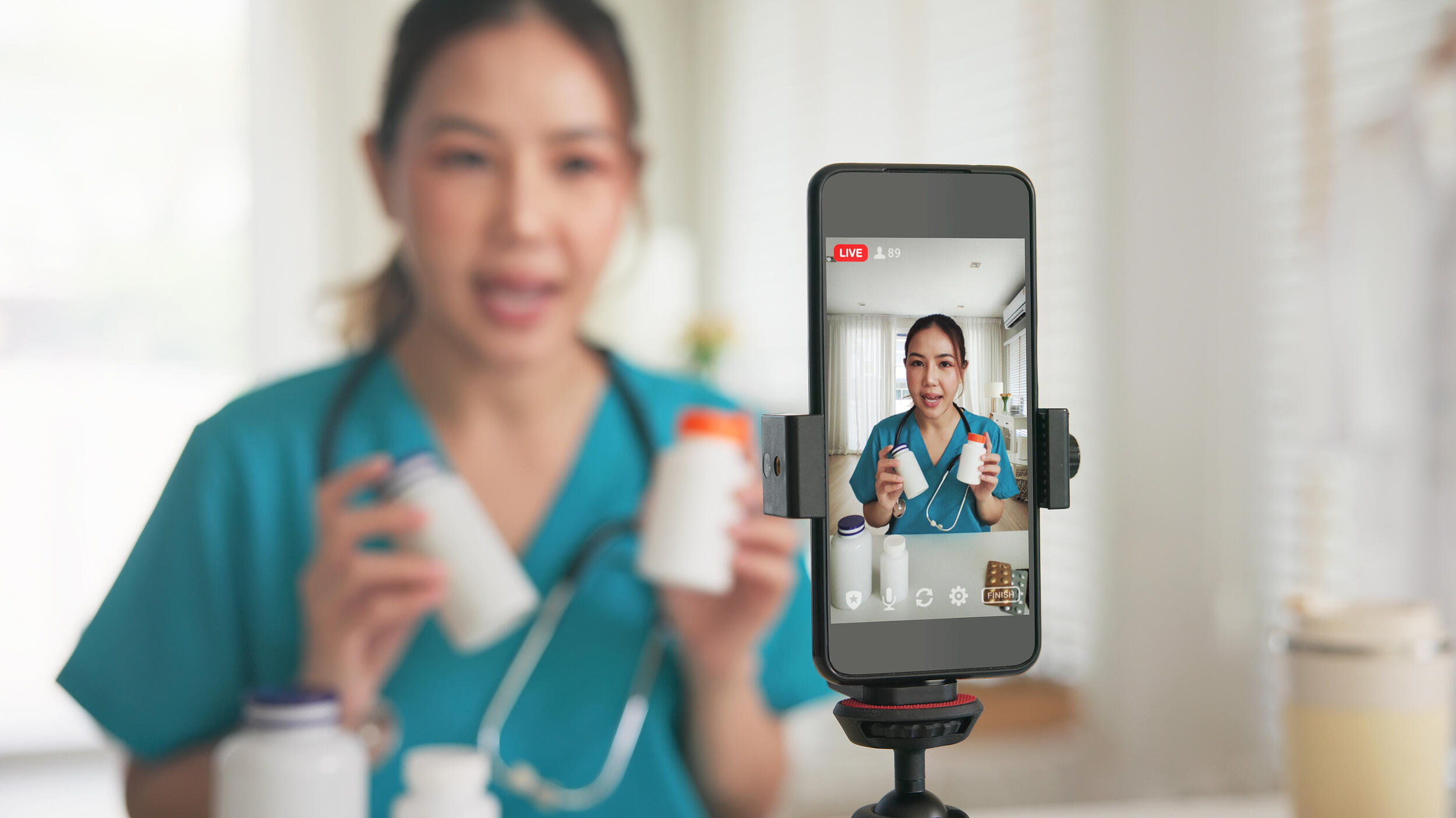 A photo of a phone on a tripod recording a woman wearing medical scrubs and a stethoscope around her neck while she talks and holds pill bottles.