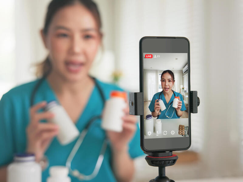 A photo of a phone on a tripod recording a woman wearing medical scrubs and a stethoscope around her neck while she talks and holds pill bottles.