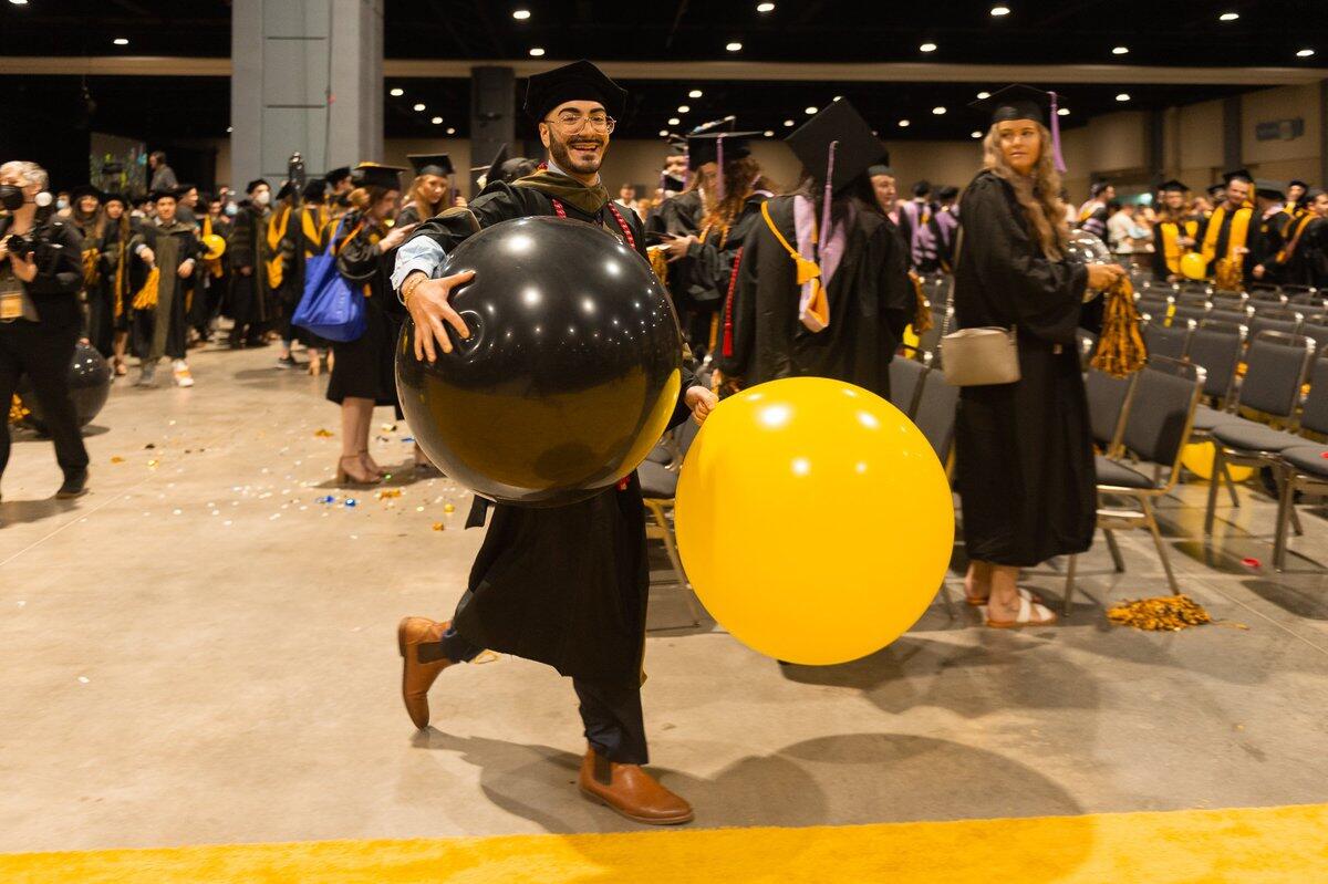 A man wearing a graduation cap and gown walking and holding two giant balloons in front of a crowd of graduating students. 