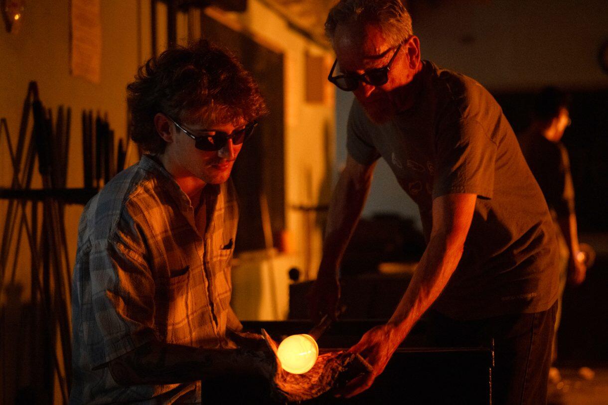 A photo of two men on either side of a glob of glowing hot glass attached to a pole.