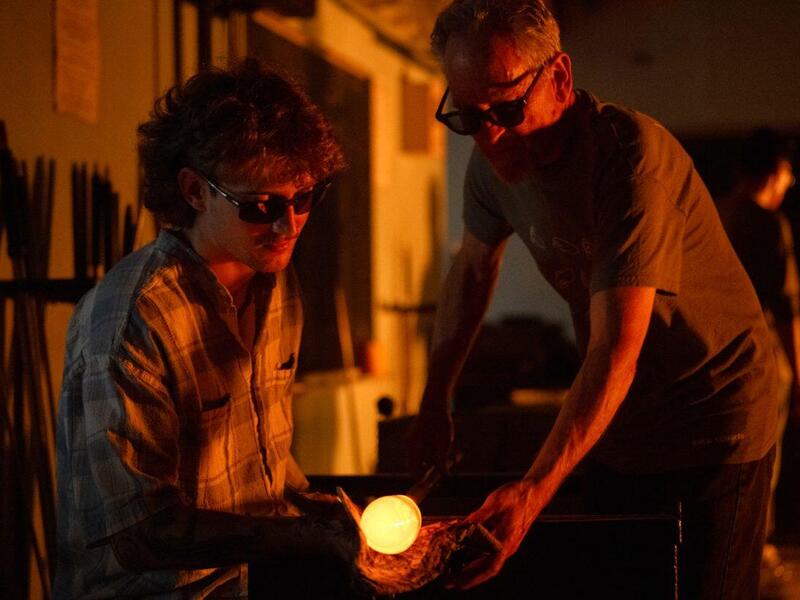 A photo of two men on either side of a glob of glowing hot glass attached to a pole.