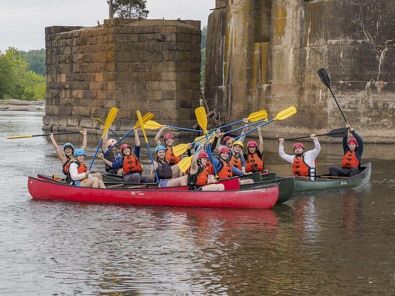 A photo of two canoes next to each other on a river. The people in the boats are cheering and raising their paddles in the air. 