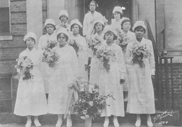 An old black and white photo of 11 women wearing nurse's uniforms holding flowers standing on a small staircase outside of the doorway to a building. 