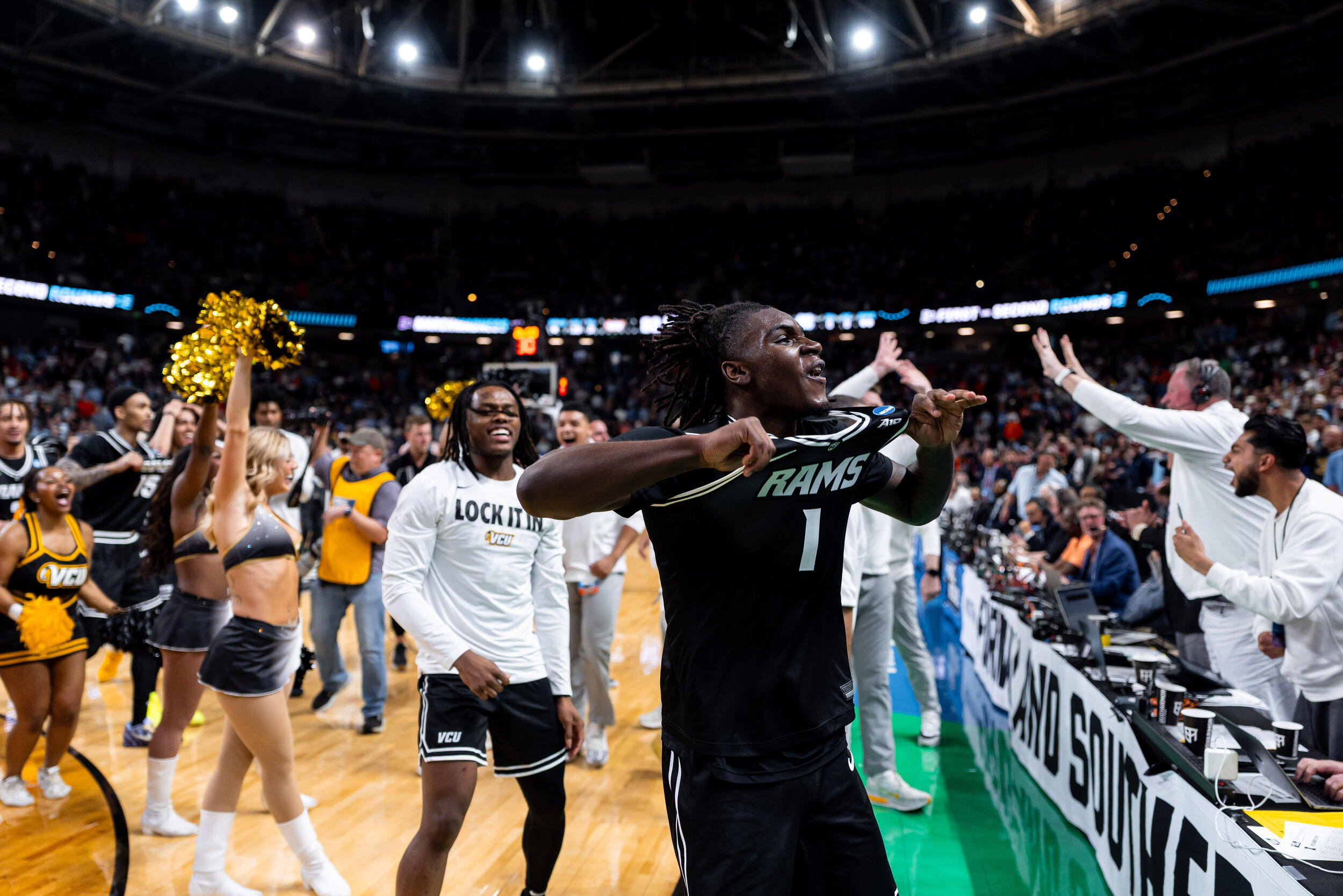 A photo of a man showing off his VCU Rams basketball jersey to a cheering crowd while standing on a court. 