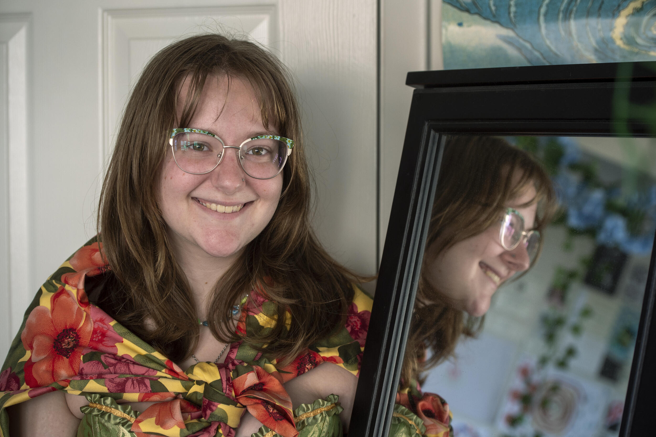 A photo of a person from the chest up sitting next to a mirror. The side of the person's face is reflected in the mirror. 