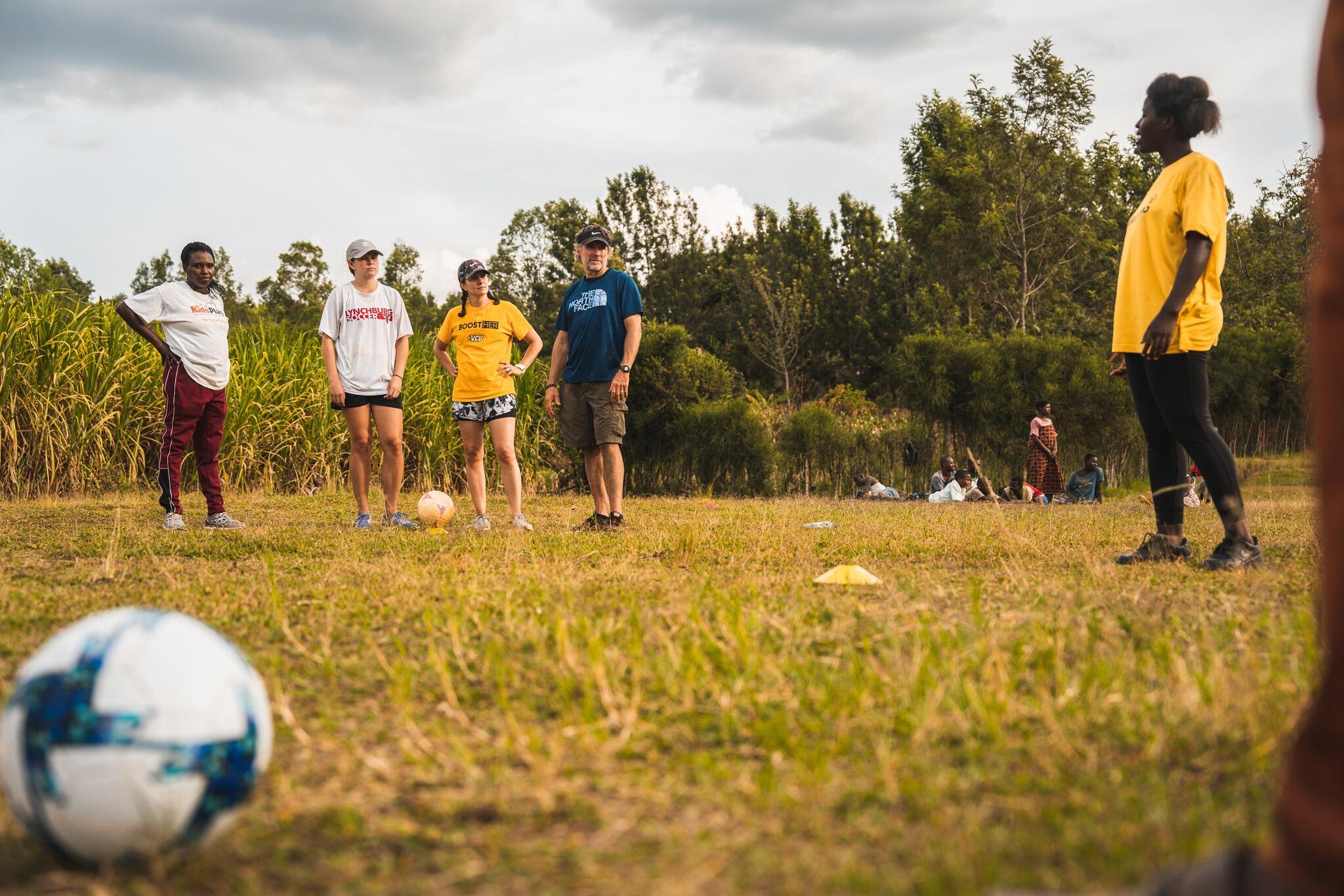 Center for Sport Leadership faculty and students stand in a soccer field along with local residents. A soccer ball is in the foreground.