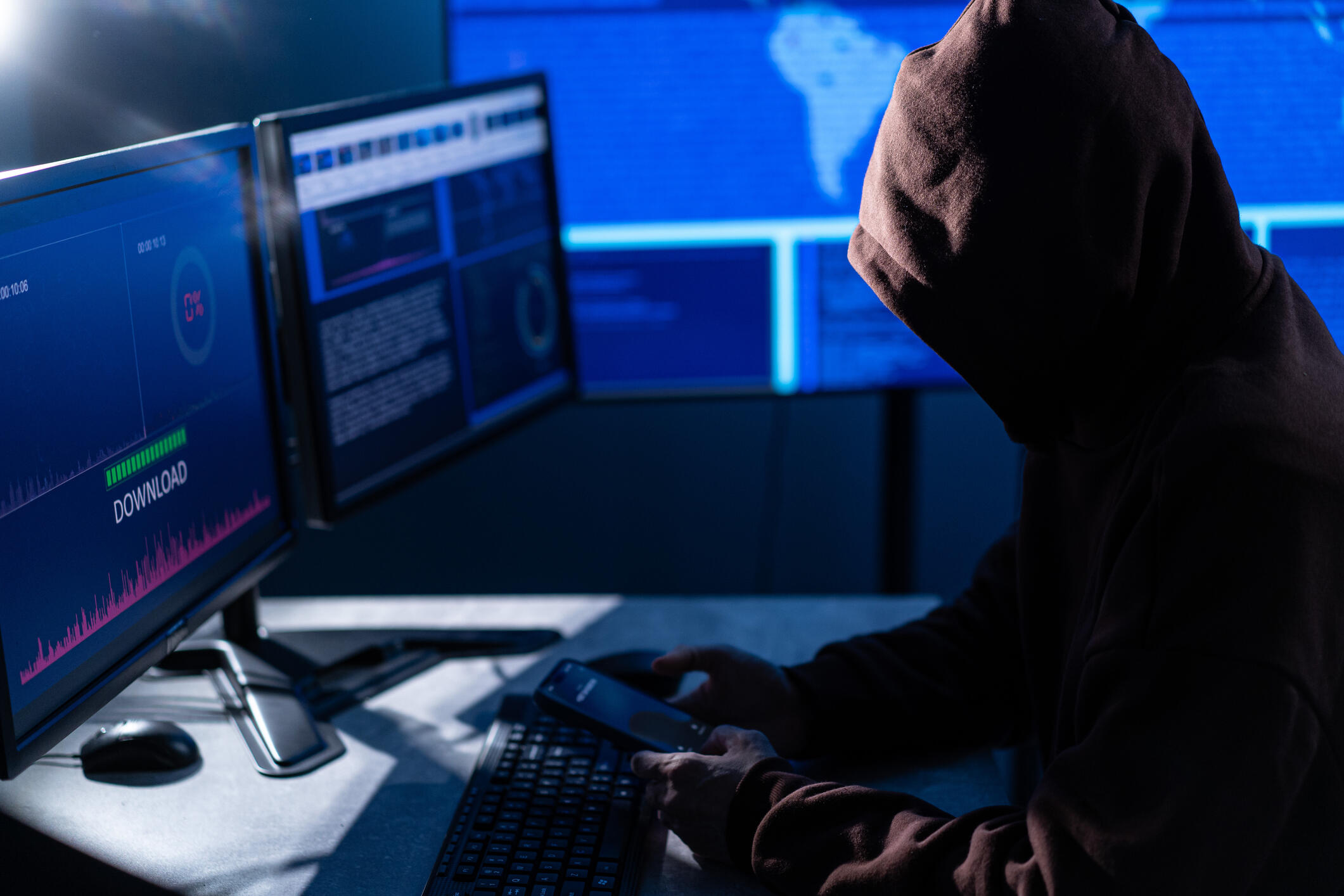 A photo of a hooded figure sitting at a desk. On the desk are two laptop screens, a compter mouse, and a keyborad.